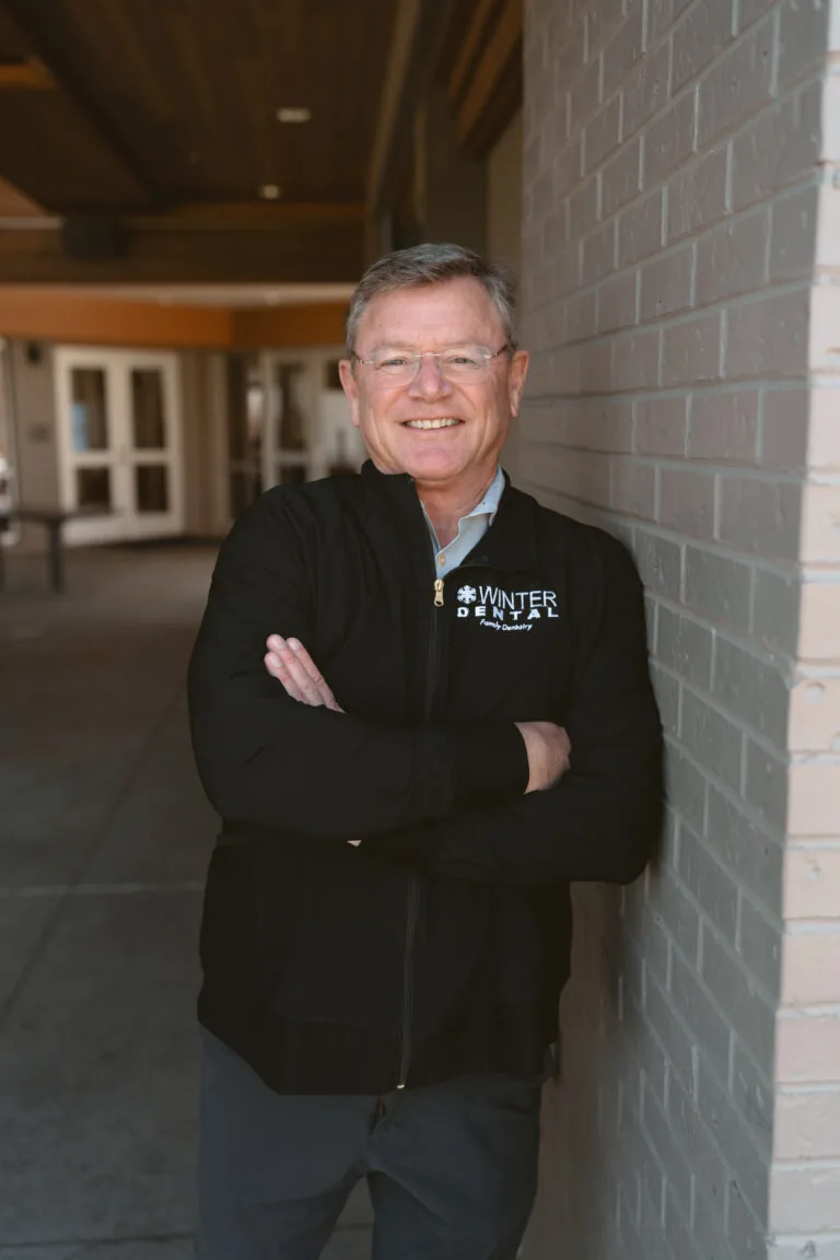 Dr. Stuart leans against a brick wall with arms crossed, wearing a black zip-up jacket embroidered with “WINTER DENTAL Family Dentistry” on the chest. The indoor hallway includes doors and windows, suggesting a casual yet professional setting, possibly in education or healthcare.