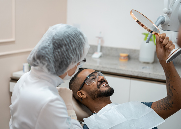 Dental clinic scene with a patient in a chair smiling at their reflection in a handheld mirror, wearing protective eyewear and a bib. A masked dental professional in a white coat sits nearby, with dental supplies visible on the countertop in the background.