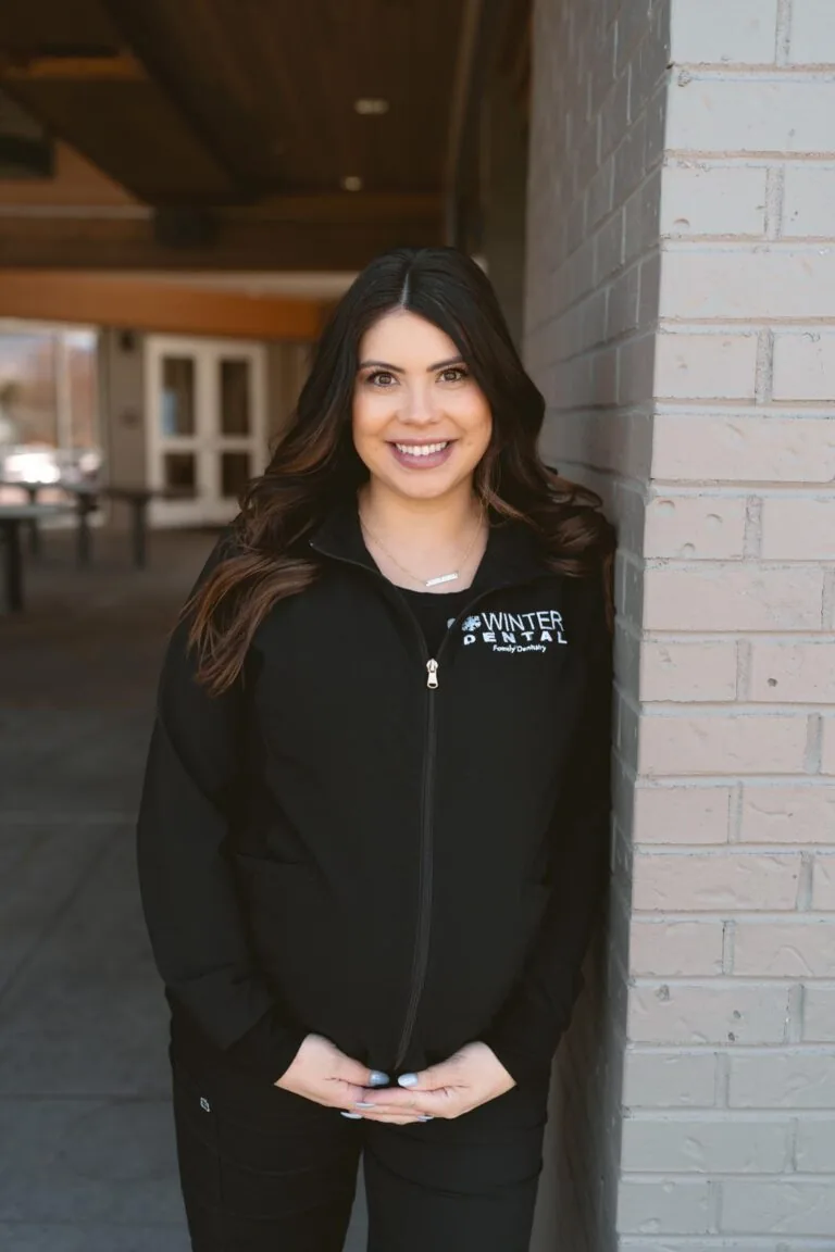 Alexis Trinidad stands in front of a brick wall wearing a black zip-up jacket labeled “WINTER DENTAL Family Dentistry.” Behind them, a building entrance with glass doors and windows suggests an educational or institutional setting.