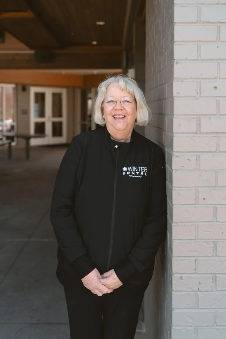 Cathy Winter- Dental Hygienist stands outdoors, leaning against a brick wall while wearing a black jacket labeled “WINTER DENTAL Family Dentistry.” The nearby building entrance and institutional surroundings evoke a professional, military-affiliated setting.