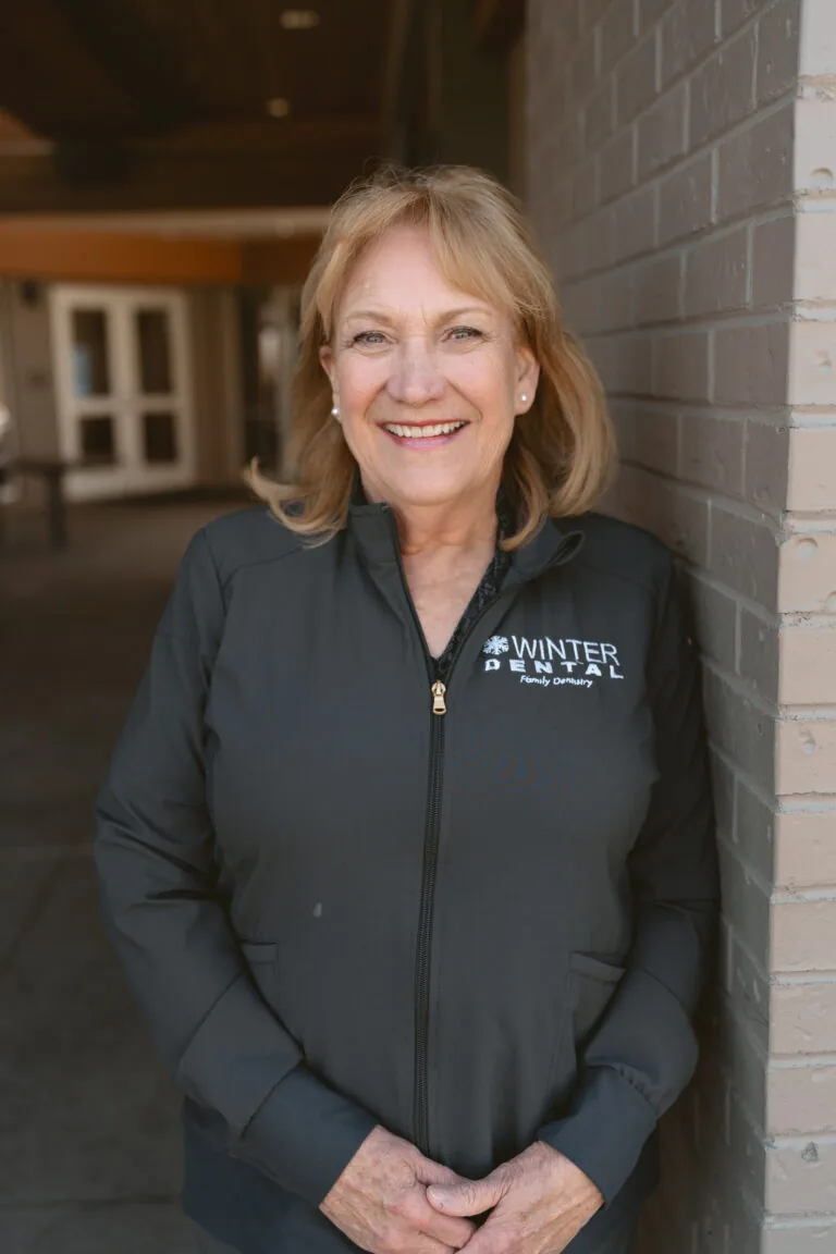 Karen Martin- Dental Hygienist stands in front of a brick wall wearing a black zip-up jacket embroidered with “WINTER DENTAL Family Dentistry” on the chest. Behind them, a building entrance and architectural elements suggest a professional dental office setting.