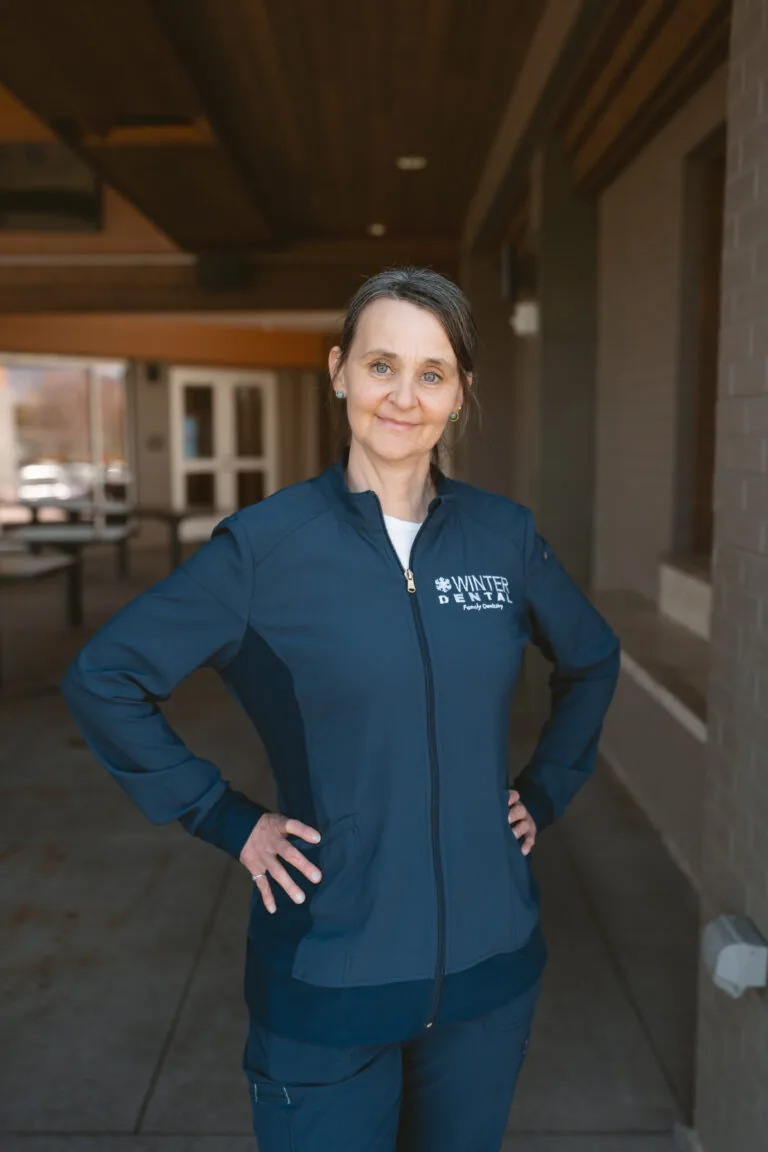 Ramona Constantin- Dental Assistant stands outdoors wearing a dark blue jacket labeled “WINTER DENTAL Family Dentistry,” hands confidently placed on the hips.