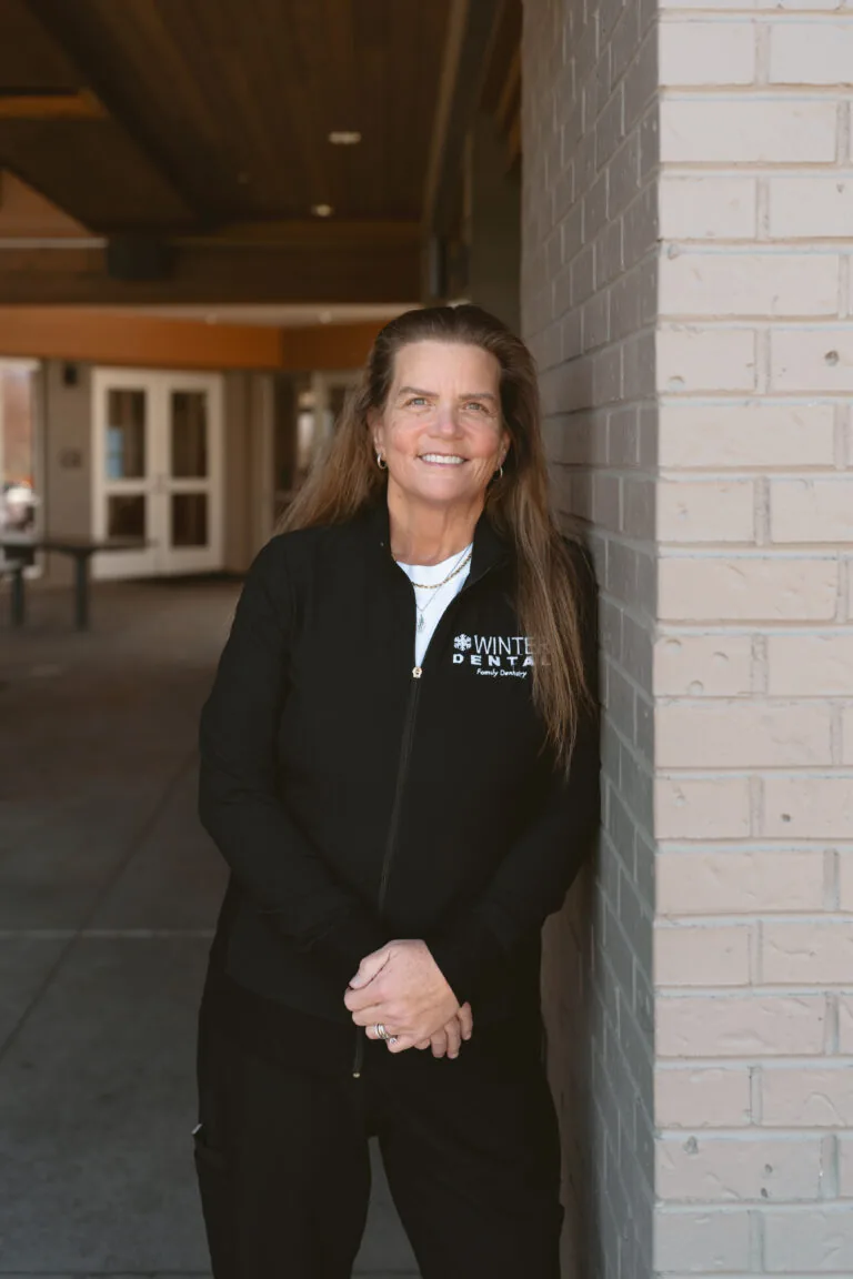 Robing Young- Dental Assistant stands in front of a brick wall wearing a black zip-up jacket with white text reading “WINTER DENTAL Family Dentistry.” Behind them, a building entrance with double doors and a bench suggests a professional or academic setting.