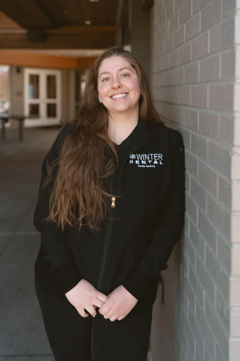 Ryan Muniz- Dental Assistant person stands in front of a brick wall wearing a black zip-up jacket embroidered with “WINTER DENTAL Family Dentistry” on the chest. The setting includes a hallway with doors and windows, suggesting affiliation with a performance group known for precision and artistry.
