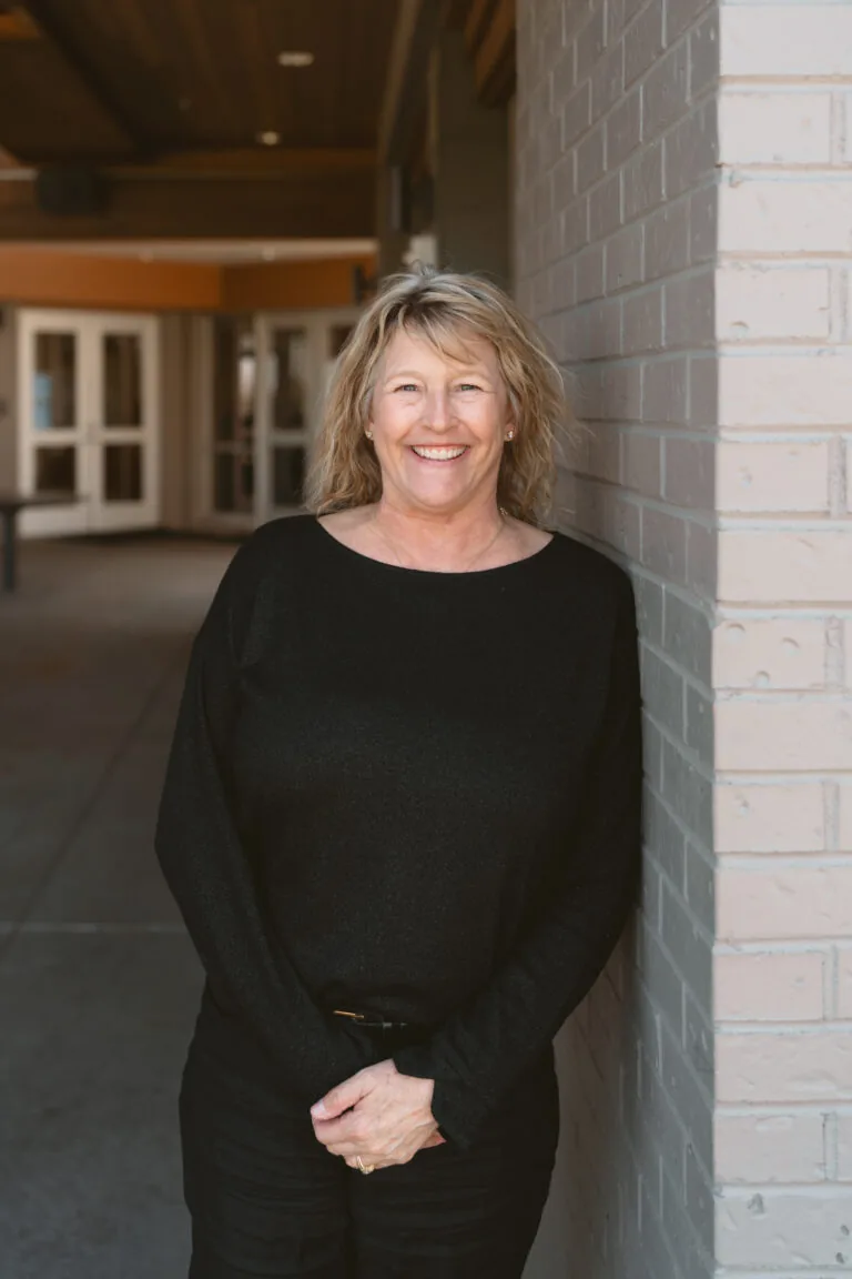 Sarah Winter- Accounting stands against a brick wall wearing a black long-sleeve shirt and black pants. The setting includes a covered building entrance with glass doors and ceiling lights, conveying a clean and professional atmosphere.