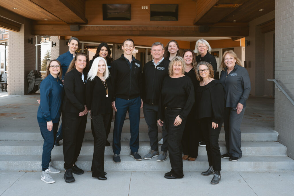 Twelve individuals stand on outdoor steps in front of a building with a wood-framed roof and large windows, most wearing black jackets labeled “WINTER DENTAL Family Dentistry.” The casual group arrangement and setting suggest a team photo outside a professional or community facility.