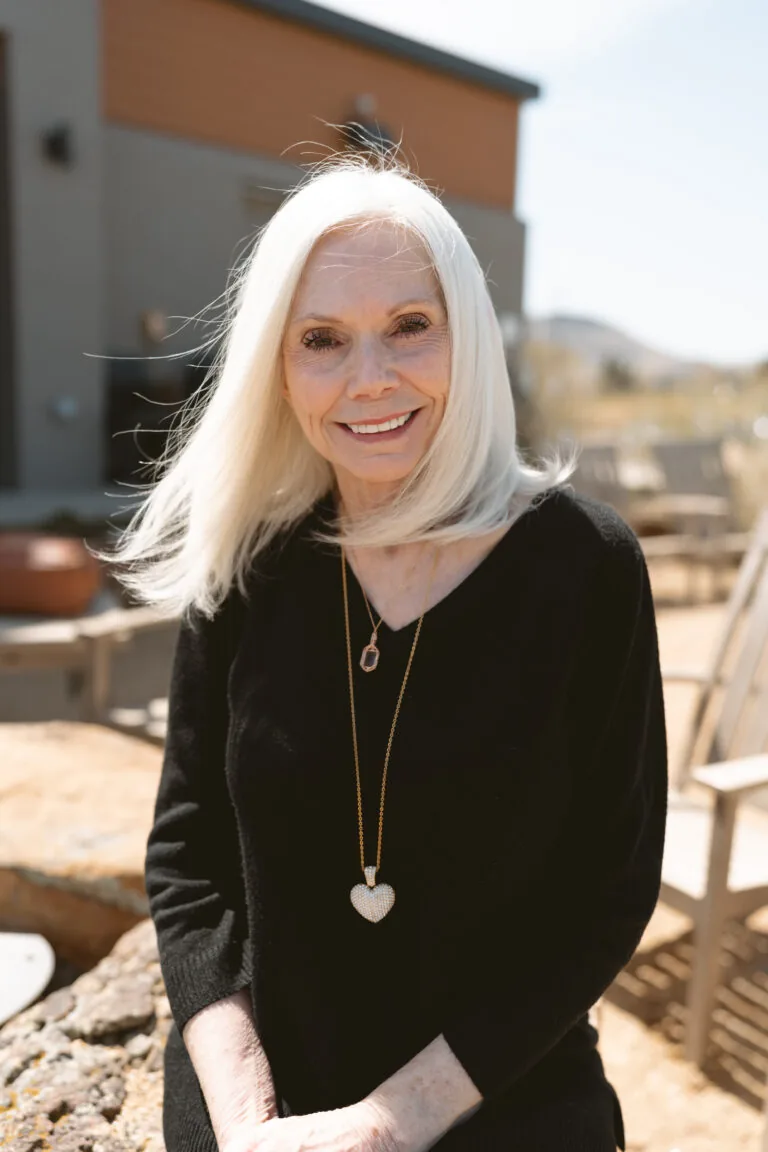 Tori Kirkland-Insurance Coordinator-Business Administration, person with long, light hair wears a black top and a necklace featuring a heart-shaped pendant, sitting outdoors. The scene includes patio furniture and part of a building, creating a relaxed and welcoming atmosphere.